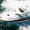 Aerial view of a Beneteau Flyer 12 yacht cruising at speed with its sliding sunroof open, showing the helmsman at the wheel, a teak swim platform, and a distinctive wood-accented hull cutting through the waves.