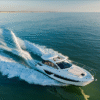 Aerial view of a Beneteau Gran Turismo series yacht cruising at speed with its sliding sunroof partially open, featuring a sleek white hull, spacious foredeck sunpad, and leaving a wide wake over calm blue-green water.