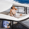 Woman sitting in the forward lounge area of a Jeanneau Leader 46 yacht, relaxing under the open sliding sunroof while the boat cruises, with the helmsman steering below.