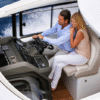 Couple seated together at the helm of a Jeanneau Leader 46 yacht, cruising with the sliding sunroof open, showing the modern dashboard and teak flooring beneath their feet.