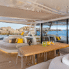 Aft deck of a Lagoon Sixty 7 yacht with a wooden dining table, curved sofas, and decorative fruit bowls, featuring a companionway hatch in the background providing access to the galley, with rocky coastline views beyond.