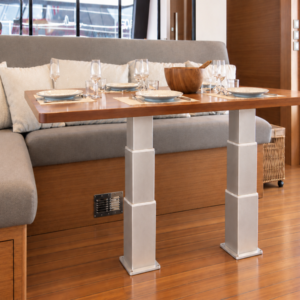 Electric table lift kit NL2388-PAIRE-B installed in a yacht saloon on a wooden floor - two synchronized 3-stage rectangular aluminium legs supporting a varnished wood dining table in front of a U-shaped bench